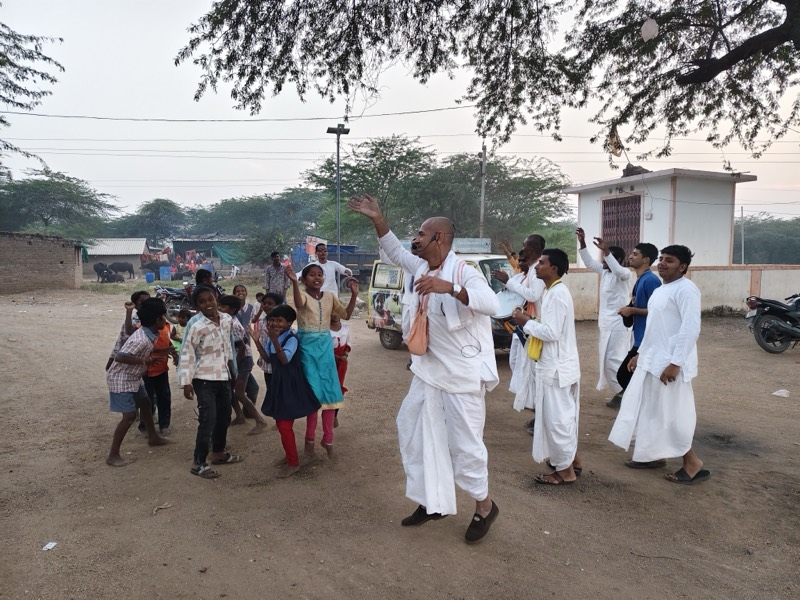 Children holding up packets of prasadam with two devotees outside a village hut
