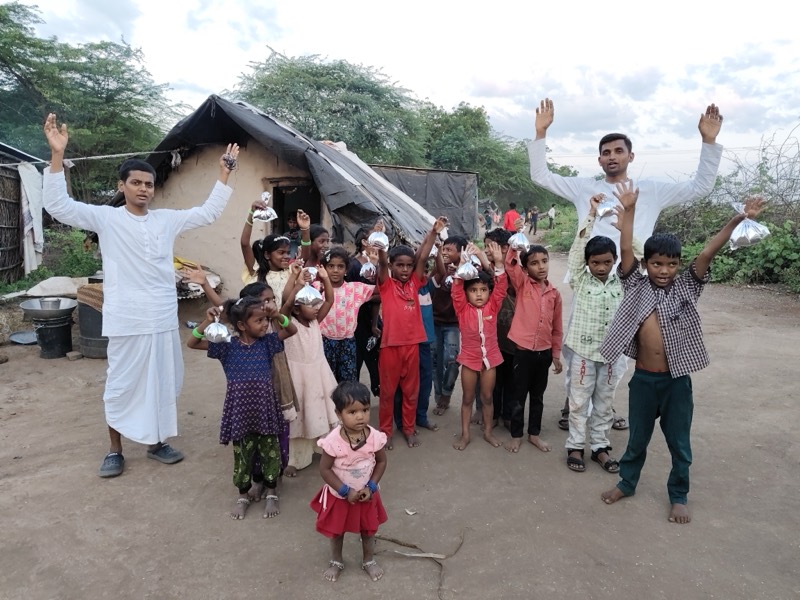 Villagers and children raising their hands in kirtan beside the prasadam van in a decorated lane