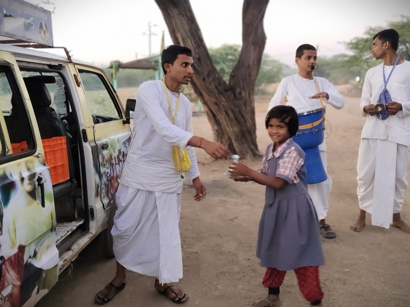 A devotee handing prasadam to a schoolgirl while another plays the mridanga